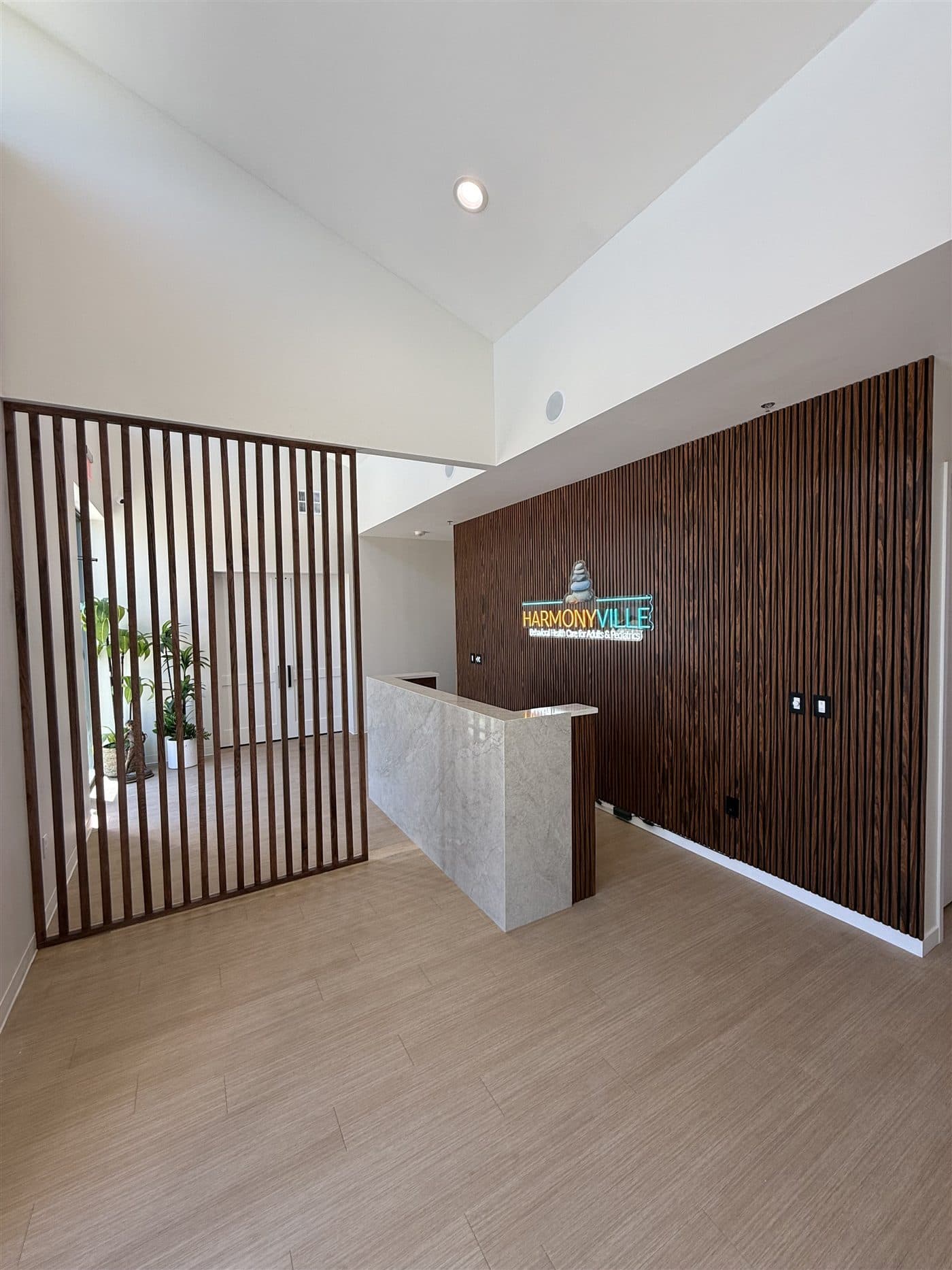 Completed Harmonyville office reception build-out with double-height slat feature wall, illuminated brand logo, custom marble reception desk, and wood slat privacy divider.