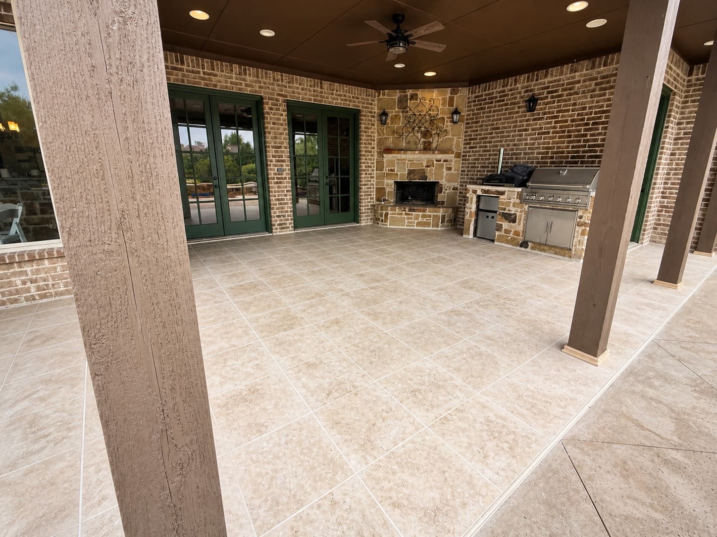 Covered patio after renovation — luxury travertine-look porcelain tile floor, refinished stone fireplace, painted column wraps, and built-in outdoor kitchen with stainless grill.