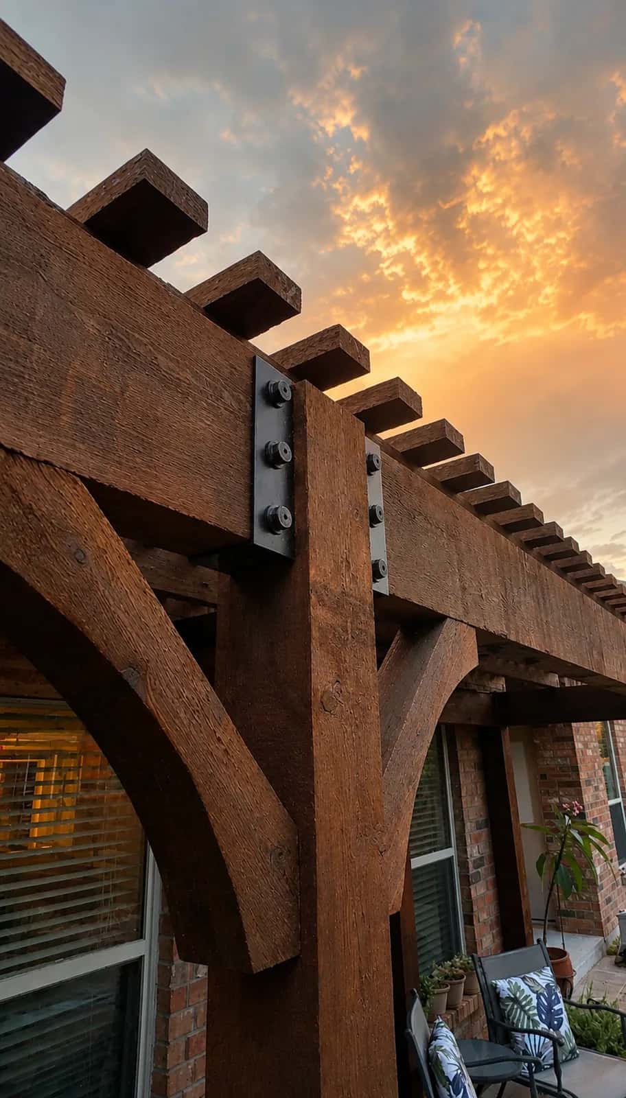 Stained cedar pergola by i30 Builders at sunset, with curved knee braces and black steel plates bolted at the post-to-beam joint.