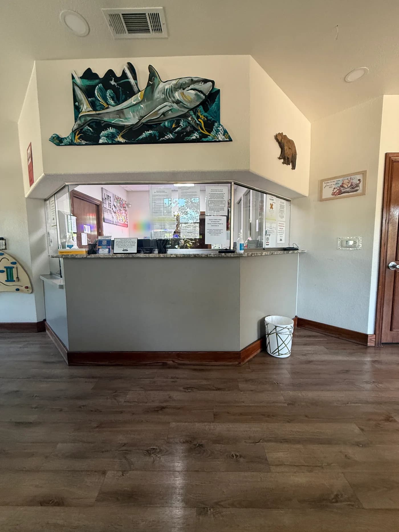 Medical office front desk before the rebuild: a painted counter with a granite top under a full glass partition covered in taped policy notices, with dated stained-wood base trim.