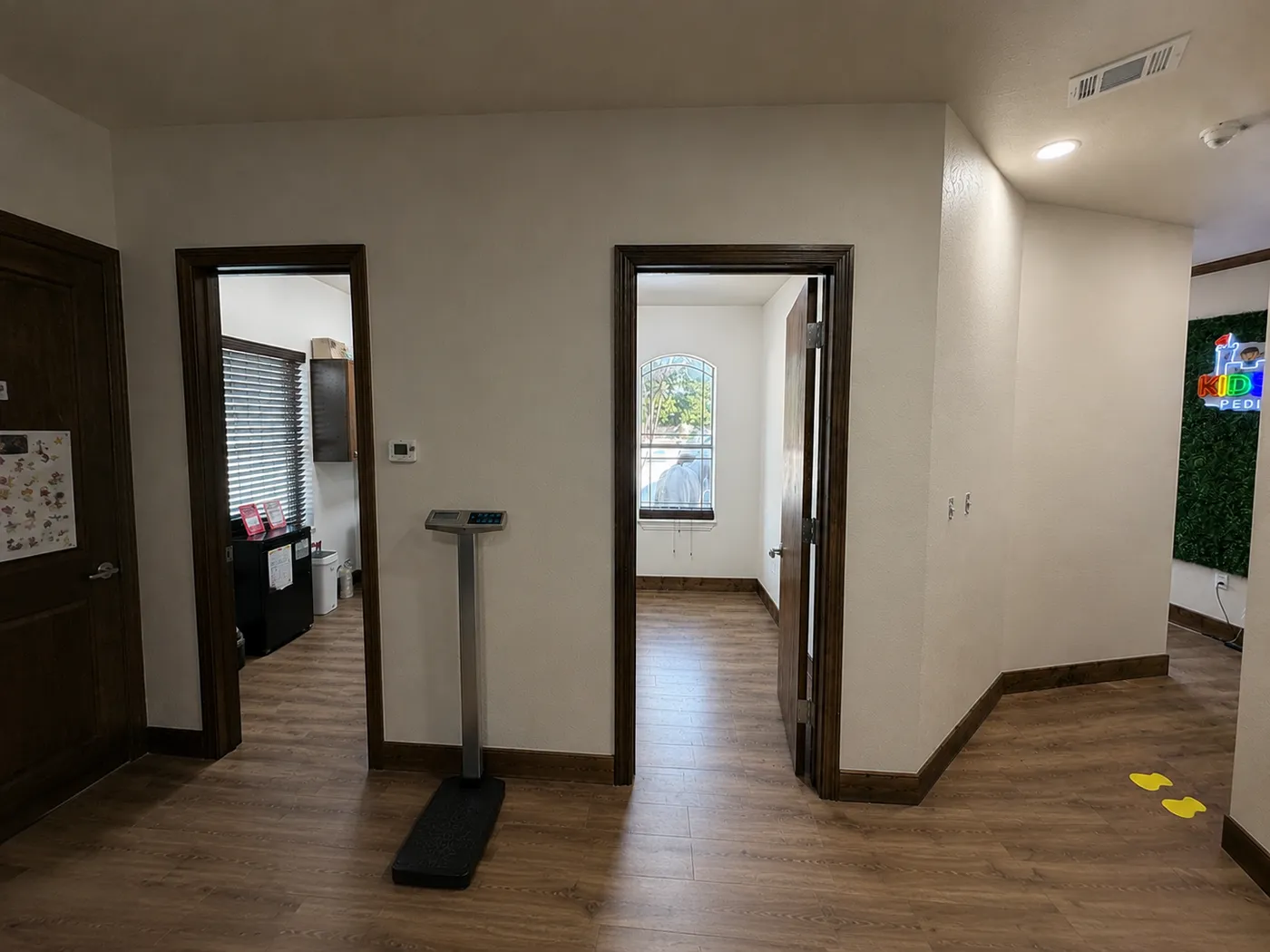 The finished clinic corridor: exam rooms off a wood-look plank hallway, stained trim and doors, a height and weight station, and a lit signage wall at the end.
