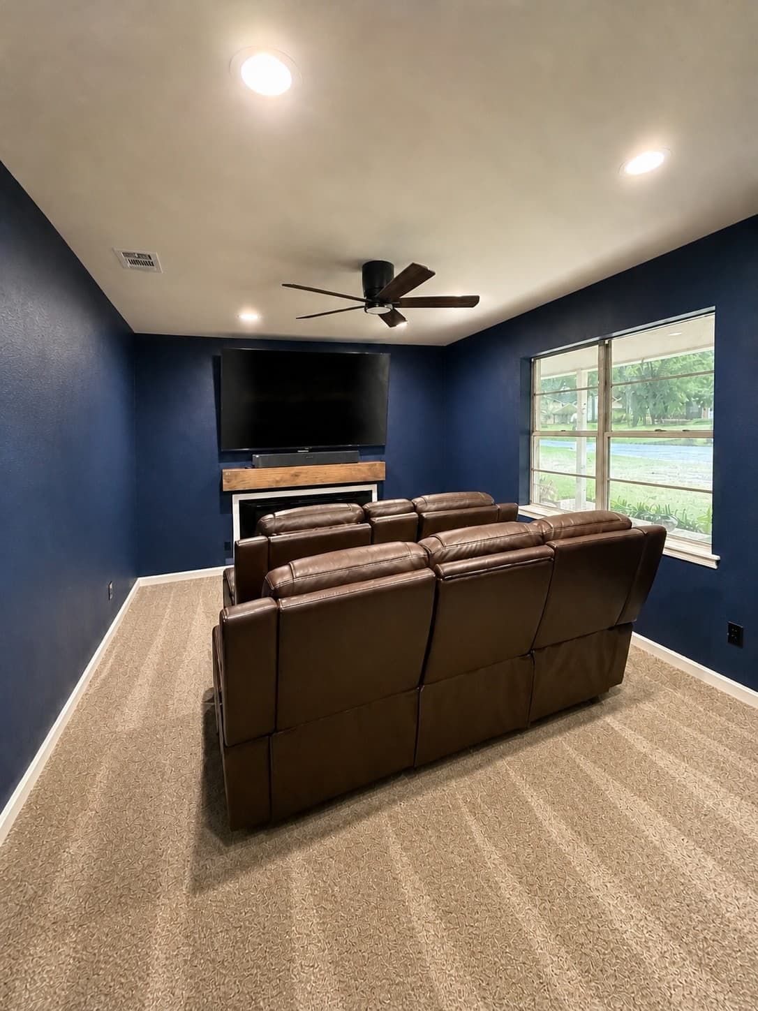 Completed media room, navy accent walls, mounted display with reclaimed-wood mantel, leather theater recliners, and new striped low-pile carpet.
