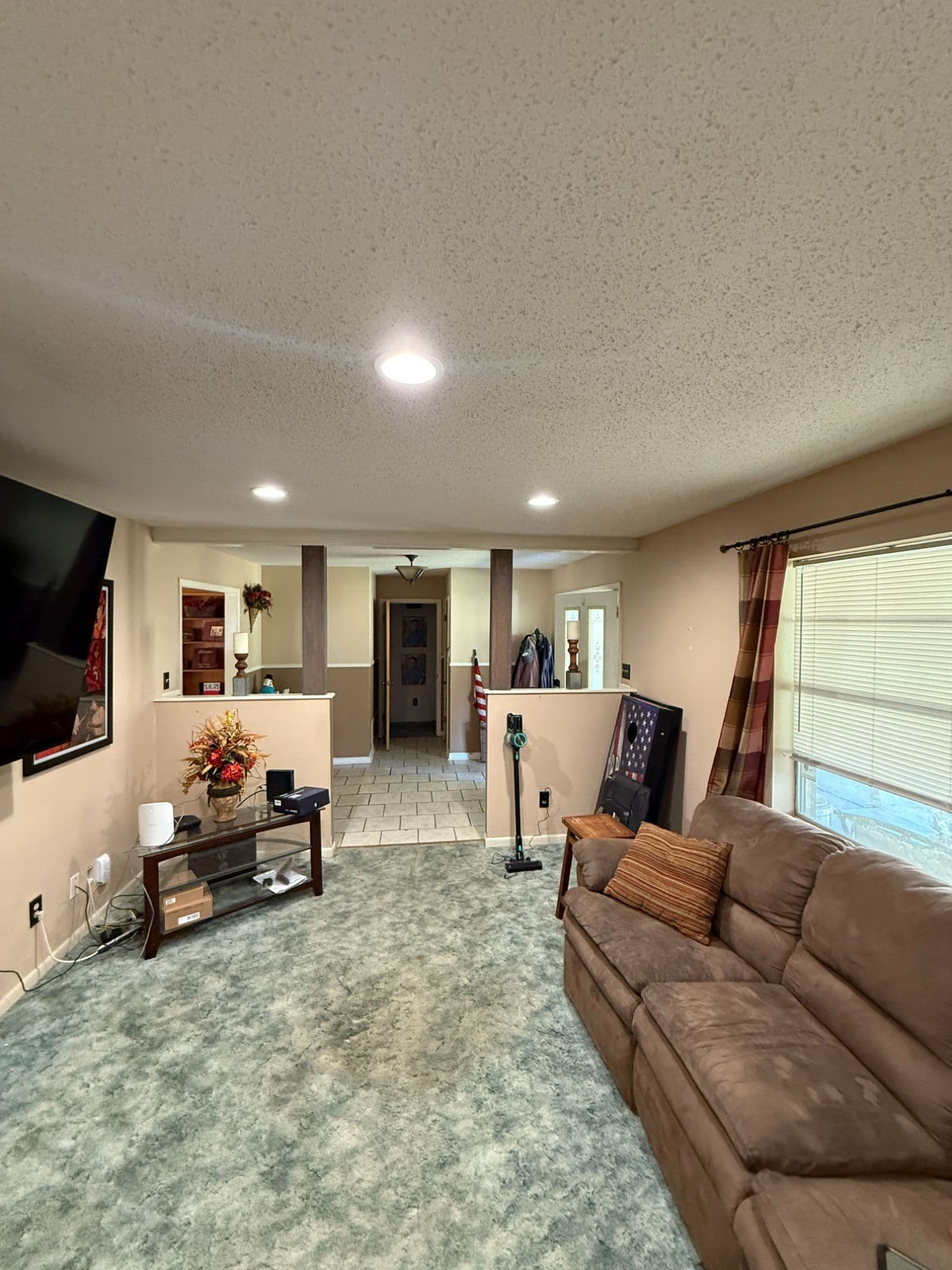 Living room before media-room conversion, beige walls, popcorn-textured ceiling, dated teal carpet, and mismatched furniture.